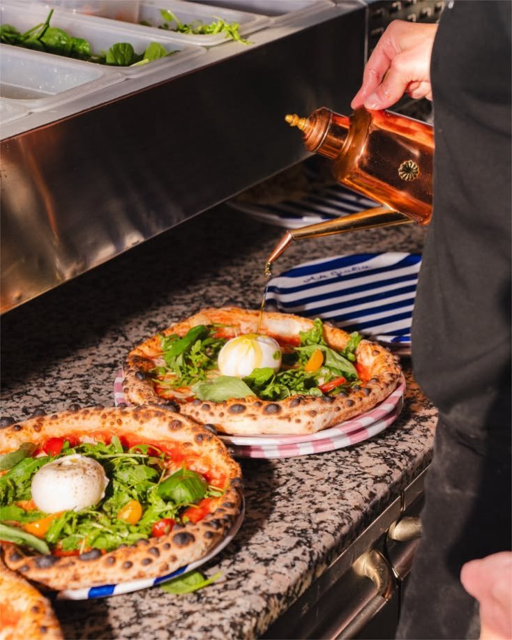 Zwei Pizzen mit Burrata, Rucola und Kirschtomaten auf der Arbeitsplatte, Olivenöl wird aus einer Kupferkanne darübergegossen