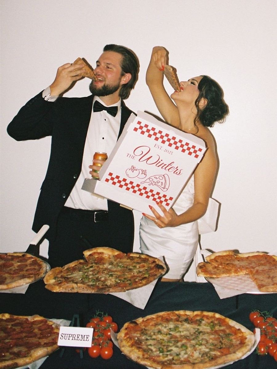 Brautpaar in Abendgarderobe isst Pizza an einem Tisch mit mehreren belegten Pizzen